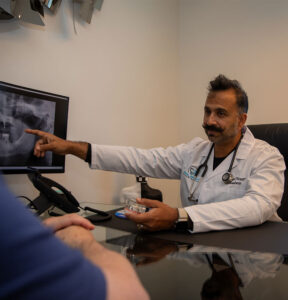 Dr. Ash Kaushesh pointing to a panoramic dental X-ray on a monitor while holding a dental implant model during a patient consultation.
