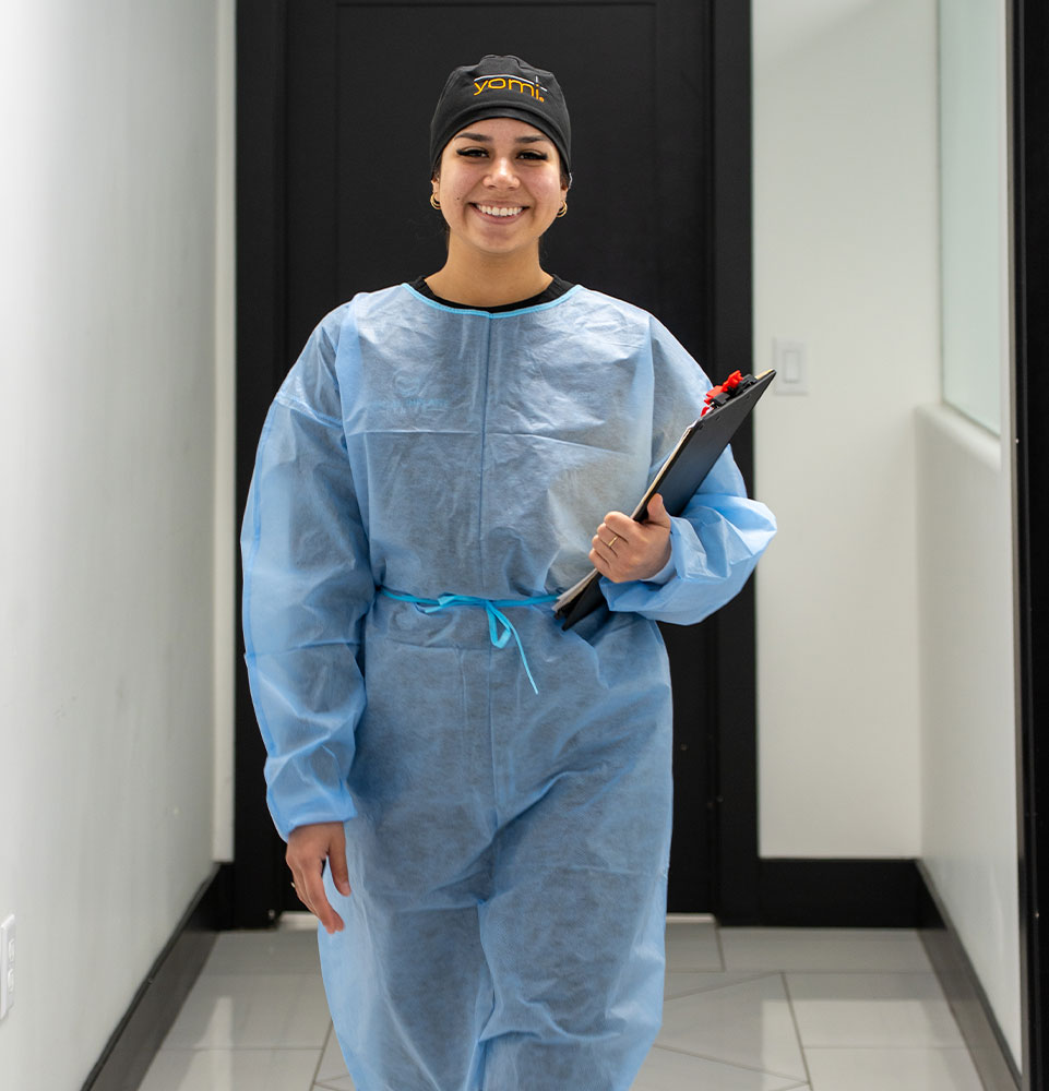 Hailey smiling in the dental office hallway, wearing a blue protective gown and a Yomi cap while holding a clipboard.